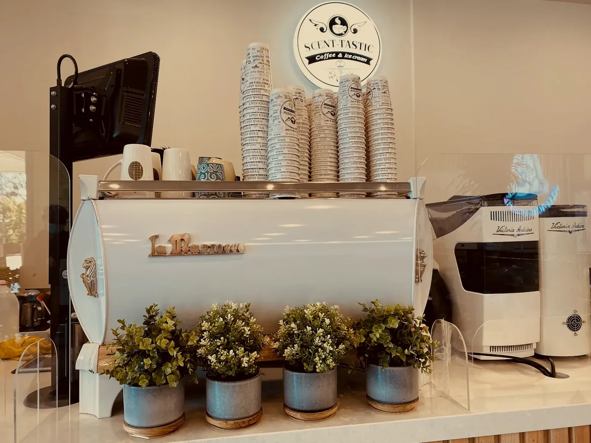 The white La Marzocco under the Scent-tastic sign, with a row of potted plants